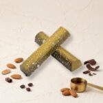 A 300x300 square professional food photograph featuring two cylindrical gourmet Amaretto truffle bars. The bars have a unique, mossy-green and gold-dusted finish with white speckles, arranged in an "X" shape in the center. The word "AMARETTO" is printed in a clean, black serif font directly below the bars. They rest on a light, textured cream surface. Artfully scattered around the base are whole almonds, roasted coffee beans, and delicate dark chocolate curls. In the bottom right, a small gold measuring scoop is filled with a golden amber liquid, suggesting Amaretto liqueur. The lighting is soft and elegant, creating a high-end, artisanal aesthetic.