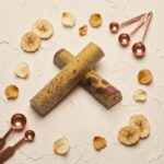 A 300x300 square professional food photograph of two cylindrical gourmet truffle bars. The bars are a light yellow-green color with heavy, irregular speckling and patterns in deep maroon and brown, resembling a ripe banana peel. They are arranged in an "X" shape on a heavily textured, off-white cream plaster surface. Scatted around the bars are artisanal accents including dried banana slices, small dried rose petals, and three polished copper-toned measuring spoons in the upper right. The text "BANANA RUM" is centered at the bottom in a bold, dark brown serif font. The lighting is soft and warm, highlighting the unique textures and curated aesthetic.