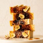 A 300x300 square professional food photograph featuring a vertical stack of five thick Bananas Foster fudge slices. Each slice has a pale yellow base topped with a rich, dark brown caramelized sugar crust and is garnished with crunchy dried banana chips. The stack sits on a smooth off-white pedestal. At the base, a small rose-gold spoon holds a dried banana slice, accompanied by scattered grains and golden droplets of caramel sauce. The background is a soft, textured cream wall, creating a warm and elegant aesthetic.