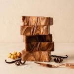 A vertical stack of five handcrafted fudge blocks with rich, distinct swirls of deep caramel brown and lighter milk chocolate forms the central focus. The blocks are precisely stacked, highlighting the intricate marbled patterns. At the base of the stack, to the left, are four whole, peeled hazelnuts and two long, dark vanilla beans. In the lower-right foreground, there are two delicate chocolate curls and a finely-crafted, rose-gold colored dessert spatula with a copper handle resting nearby. The entire presentation is set against a seamless, light cream-colored textured background, creating a warm, elegant product showcase.