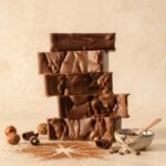 A 300x300 square professional food photograph featuring a stack of five thick, decadent Belgian chocolate fudge slices. The fudge has a glossy, rippled texture on the sides, showcasing its creamy consistency. The stack is centered on a light beige surface against a textured cream background. At the base, the scene is decorated with cocoa powder dusted in a starburst pattern, dark chocolate curls, and whole hazelnuts. To the right, a small hand-painted ceramic bowl contains cocoa nibs with a rose-gold spoon resting inside. The overall aesthetic is warm, sophisticated, and artisanal.