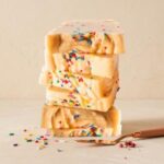 A 300x300 square professional food photograph of five thick, artisanal fudge slices stacked high. The fudge is a pale, creamy off-white color, generously filled and topped with vibrant multi-colored rainbow sprinkles. The stack sits on a smooth cream-colored surface against a textured, soft cream background. To the left, several colorful sprinkles are artfully scattered. In the foreground, a rose-gold dessert spatula rests on the surface alongside a small cluster of pink peppercorns. The lighting is soft and warm, creating a clean and festive high-end presentation.