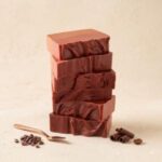 A close-up, central photograph of six rectangular blocks of rich, dark chocolate coffee fudge stacked vertically. Each block has a smooth top and sides, with a decorative wavy swirl patterned into the chocolate. The stack is centered against a warm, light textured beige background. Surrounding the base are elements that highlight the ingredients and craft: a cluster of whole roasted coffee beans to the lower left, a polished copper tasting spoon resting adjacent to them, and two delicate dark chocolate curls with a single coffee bean on the surface to the lower right. The lighting is soft and directional, emphasizing the texture and rich