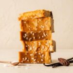 A 300x300 square artisanal food photograph featuring a tall, slightly uneven vertical stack of six rectangular salted caramel fudge blocks. Each piece is a rich, warm golden-caramel color and is generously topped with visible coarse sea salt flakes. The stack is centered on a textured, light cream-colored surface. At the base, a small copper tasting spoon holds a few salt crystals, and two whole, dark dried vanilla bean pods rest alongside a whole roasted cocoa bean. The background is a soft, warm cream wall, creating a sophisticated and appetizing presentation.