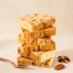 A 300x300 square professional food photograph featuring a tall, slightly offset stack of six thick butter pecan fudge slices. The fudge is a warm, creamy tan color, densely packed with visible pieces of roasted pecans and topped with a light dusting of pecan crumbles. The stack is centered on a textured, off-white surface against a soft cream background. In the lower-right foreground, a single whole pecan sits next to a small cluster of raisins and granola. A rose-gold dessert spatula handle enters the frame from the bottom left, completing the high-end, artisanal presentation.