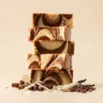 A 300x300 square image featuring a stack of five thick, rectangular fudge slices with a beautiful swirl of dark chocolate and creamy cheesecake flavors. The marbled pattern creates elegant, flowing waves of brown and off-white. The stack is set against a soft, warm cream background. At the base, the scene is decorated with chocolate curls, white chocolate shavings, whole coffee beans, and vanilla beans dipped in white chocolate. A delicate gold wire swirl adds a final artisanal touch to the professional food photography.