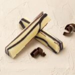 A 300x300 square professional food photograph featuring two cylindrical gourmet Chocolate Cheesecake truffle bars. The bars are arranged in an "X" shape and feature a smooth, creamy ivory-white finish decorated with elegant, thick dark chocolate marble swirls. The text "CHOCOLATE CHEESECAKE" is centered at the bottom in a sophisticated, copper-toned serif font. The bars rest on a heavily textured, light cream plaster surface. The scene is artfully decorated with delicate dark chocolate curls scattered around the base. The lighting is soft and bright, creating a clean, premium, and artisanal aesthetic.
