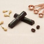 A high-resolution product photograph from an overhead angle, featuring two crossed cylindrical gourmet chocolate truffle bars on a cream-colored textured plaster surface. The bars are made of dark chocolate with internal white and green swirls, centrally placed on the plaster surface. Surrounding them are the same carefully curated aesthetic items from image_280.png: scattered dried jasmine flower buds on the left, a single roasted coffee bean and jasmine bud between the bars, and the two copper measuring spoons in the upper right. The gold serif text 'CHOCOLATE MINT' is retained below the bars. The scene is well-lit with soft, directional light, highlighting the textures of the bars, props, and plaster. The entire composition is balanced and has a curated, sophisticated feel.