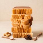 A 300x300 square professional food photograph featuring a tall stack of six thick fudge slices. Each slice has a distinct two-layer composition: a rich, dark chocolate base topped with a smooth, creamy peanut butter layer. The stack is centered on a textured, light cream-colored surface. In the lower-left foreground, a small cluster of whole roasted peanuts sits next to a rose-gold measuring spoon filled with coarse sea salt. To the right, three delicate dark chocolate curls and a rose-gold dessert spatula complete the artisanal scene. The background is a soft, warm cream, emphasizing the fudge's rich, decadent textures.