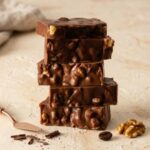 A 300x300 square professional food photograph featuring a tall, slightly offset stack of five thick, rectangular chocolate walnut fudge slices. The fudge is a rich, dark brown color with a bumpy, highly-textured surface packed with visible large walnut pieces. It sits centered on a warm, textured cream-colored surface. In the foreground, there is a small cluster of whole roasted walnuts to the lower-right and a single whole coffee bean to the lower-left. A small, elegant, rose-gold colored dessert spatula handle enters the frame from the bottom left. The entire background is a soft, warm cream, giving the image a clean and high-end aesthetic. The lighting is soft and directional, highlighting the rich textures of the fudge and scattered ingredients.