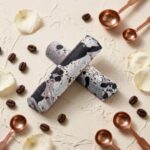 A close-up, high-angle photograph of two cylindrical gourmet truffle bars. The bars feature a distinct, speckled white and dark chocolate finish, resembling the Cookies 'N' Cream flavor or dalmatian spots. They are arranged in an "X" shape on a heavily textured, off-white cream colored stone or plaster surface. Scattered around the bars are whole roasted coffee beans, a few whole dried white rose petals, and four polished copper-toned measuring spoons. The lighting is soft and natural, emphasizing the textures and curated aesthetic.
