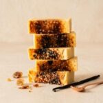 A 300x300 square professional food photograph of five thick, rectangular fudge blocks stacked vertically. The fudge is a pale, creamy custard color, featuring a dramatic, crackled dark brown caramelized sugar topping that mimics a classic crème brûlée. The stack is centered on a textured cream-colored surface. To the left, several shards of amber-colored caramelized sugar glass are artfully scattered. On the right, two long, dark vanilla bean pods rest beside a small, polished rose-gold dessert spoon. The background is a soft, warm cream, creating a sophisticated and indulgent artisan display.