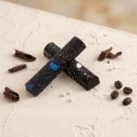 A 300x300 square professional food photograph of two cylindrical gourmet Dark Almond Viennese truffle bars. The bars are arranged in an "X" shape and feature a deep dark chocolate base, heavily speckled with fine white dots and occasional splashes of vibrant blue. The text "DARK ALMOND VIENNESE" is centered at the bottom in a bold, dark brown serif font. The bars rest on a heavily textured, light cream plaster surface. Surrounding the bars are artisanal accents including dark chocolate curls, roasted coffee beans, and small white decorative pearls. The lighting is soft and sophisticated, highlighting the unique textures and colors for a high-end, curated look.
