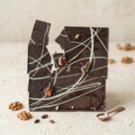 A 300x300 square professional food photograph featuring a large, dark Belgian chocolate bar that has been broken into several jagged, artistic pieces. The surface of the chocolate is decorated with thin, abstract white chocolate swirls and small pieces of embedded orange-bourbon soaked pecans. The chocolate rests on a light, textured cream-colored surface. In the foreground, several whole walnuts, roasted cocoa nibs, and a small copper measuring spoon filled with coarse sea salt are artfully scattered. The background is a minimalist, warm cream, creating a sophisticated and rustic aesthetic. Soft, directional lighting highlights the rich, dark texture of the broken chocolate and the delicate white swirls.