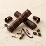 A 300x300 square professional food photograph of two cylindrical gourmet truffle bars. The bars are made of rich dark chocolate and feature elegant, diagonal peanut butter-colored swirls. They are arranged in an "X" shape on a textured, off-white cream surface. Surrounding the bars are artfully placed dark chocolate curls, a single dark vanilla bean pod, and several roasted coffee beans. The lighting is soft and warm, highlighting the glossy finish of the chocolate and the artisanal, high-end presentation.