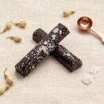 A 300x300 square professional food photograph featuring two cylindrical gourmet "Dark Coconut" truffle bars. The bars are made of rich dark chocolate and are heavily decorated with fine white speckles and larger white splatters, evoking the look of shredded coconut or falling snow. They are arranged in an "X" shape on a soft, cream-colored woven linen surface. The scene is elegantly accessorized with small dried jasmine flower buds, large crystals of translucent sea salt, and a single copper-toned measuring spoon in the corner. The text "DARK COCONUT" is centered at the bottom in a bold, dark chocolate-toned serif font. The lighting is soft and natural, creating a clean, high-end, and artisanal aesthetic.