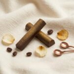 A 300x300 square professional food photograph featuring two cylindrical gourmet "Dark Espresso" truffle bars. The bars are made of rich dark chocolate and are heavily dusted with edible bronze-gold luster dust, giving them a metallic shimmer. They are arranged in an "X" shape on a soft, cream-colored woven linen surface. The scene is elegantly accessorized with whole roasted dark coffee beans, small dried natural flower petals, and two tiny, copper-toned measuring spoons in the upper right. The text "DARK ESPRESSO" is centered at the bottom in a bold, dark brown serif font. The lighting is soft and natural, highlighting the metallic dust and textures for a high-end, artisanal aesthetic.
