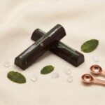 A 300x300 square professional food photograph of two cylindrical gourmet "Dark Mint" truffle bars. The bars are made of rich dark chocolate and feature an artistic top surface with a vibrant green stripe and white chocolate splatters. They are arranged in an "X" shape on a soft, cream-colored woven linen surface. The scene is elegantly accessorized with fresh green mint leaves, large translucent sugar crystals, and two tiny, copper-toned measuring spoons in the corner. The text "DARK MINT" is centered at the bottom in a bold, dark brown serif font. The lighting is soft and natural, highlighting the glossy finish and curated aesthetic.
