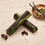 An eye-level close-up studio photograph of two dark chocolate and lime green marbled cylindrical truffle bars arranged in an "X" shape on a textured cream surface. The top bar is longer, creating a dynamic composition. In the background, to the right, a patterned copper dish holds three dark vanilla bean pods crossed over each other and a cluster of light green cardamom pods. In the left and center foreground, delicate dark chocolate curls are artfully scattered. The text "BLK BAR" is centered above the crossed truffles, and the text "DARK PISTACHIO" is centered below, both in a clean, black serif font. The lighting is soft and even, highlighting the textures of the truffles, fabric, vanilla, cardamom, and chocolate curls for an upscale, curated aesthetic.