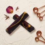 A 300x300 square professional food photograph of two cylindrical gourmet truffle bars. The bars are made of rich dark chocolate and feature elegant red and yellow swirls. They are arranged in an "X" shape on a textured, off-white cream surface. Decorative elements scattered around the bars include a dried pink and yellow rose petal, fine chocolate shavings, and five polished copper-toned measuring spoons. The lighting is soft and warm, highlighting the gloss of the chocolate and the curated, artisanal aesthetic.