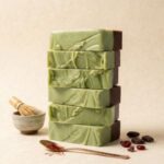 A tall, precise vertical stack of six rectangular handcrafted fudge bars forms the centerpiece against a light cream-textured background. Each bar features a thick layer of swirled, pale green matcha fudge on the left, with visible texture and a few darker flecks, and a smaller, dark brown chocolate layer on the right. The stack is centered. To its left, a small, light gray ceramic matcha bowl holds a traditional bamboo chasen (matcha whisk). Below the stack, a hammered, rose-gold colored spoon rests with a small pile of saffron threads. To the right, several loose, dark whole cacao beans and a few dried red cranberries are scattered on the surface. The entire presentation is aesthetic, with warm, soft natural lighting and a curated, artisanal feel. The square frame has clean lines and is well-balanced.