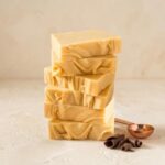 A 300x300 square professional food photograph featuring a tall stack of six rectangular French vanilla fudge blocks. The fudge is a consistent, pale buttery yellow with soft, artisanal ripples on the sides. The stack is centered on a textured, light cream surface. In the lower-right foreground, three dark chocolate curls rest near a small, polished copper-toned measuring scoop. The background is a minimalist, warm cream color, emphasizing the smooth texture and elegant simplicity of the fudge. Light, soft shadows add depth to the artisanal presentation.