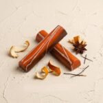A 300x300 square professional food photograph featuring two cylindrical gourmet "Fresh Orange" truffle bars. The bars have a vibrant, glossy burnt-orange finish decorated with delicate, sweeping white chocolate swirls. They are arranged in an "X" shape on a neutral, textured cream plaster surface. The scene is artistically styled with curls of dried orange zest, a whole star anise, and thin vanilla bean strands. The text "FRESH ORANGE" is centered at the bottom in a bold, dark brown serif font. The lighting is soft and natural, creating a warm and sophisticated artisanal look.