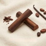 A 300x300 square professional food photograph featuring two cylindrical gourmet "Fudge Brownie" truffle bars. The bars are made of rich dark chocolate and are arranged in an "X" shape on a soft, cream-colored woven linen surface. The scene is elegantly accessorized with whole roasted coffee beans, small curls of chocolate shavings, and a single whole dried vanilla bean. The text "FUDGE BROWNIE" is centered at the bottom in a bold, dark chocolate-toned serif font. The lighting is soft and natural, creating a high-end, artisanal aesthetic.