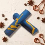 A 300x300 square professional food photograph featuring two cylindrical gourmet "Hazelnut" truffle bars. The bars are decorated with a striking artistic design of vibrant cobalt blue and bright yellow marble swirls. They are arranged in an "X" shape on a textured, off-white cream plaster surface. The scene is elegantly accessorized with scattered roasted coffee beans, whole star anise pods, and polished copper-toned measuring spoons in the corners. The text "HAZELNUT" is centered at the bottom in a bold, dark brown serif font. The lighting is soft and natural, emphasizing the bold colors and curated, high-end aesthetic.