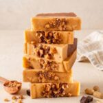 A 300x300 square professional food photograph of a tall stack of six rectangular fudge blocks. Each slice features a base of dark chocolate topped with a thick layer of creamy golden-tan toffee fudge. The front of each slice is decorated with a generous cluster of crushed toffee and chocolate bits. The stack is centered on a textured cream surface against a soft cream background. In the lower-left, a copper measuring scoop overflows with toffee crumbles. To the right, several small dark chocolate squares are artfully placed. The lighting is warm and bright, highlighting the crunchy texture of the toffee toppings.