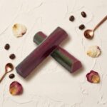 A professional food photograph featuring two crossed cylindrical gourmet truffle bars. The bars have a deep burgundy finish with subtle green undertones, presented on a cream-colored textured plaster surface. The scene is elegantly curated with scattered roasted coffee beans, small dried natural flower petals, and two tiny polished copper measuring spoons in the corners. Centered below the chocolate bars, the text "HOT MESS" is written in a sophisticated brown serif font.