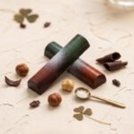 A 300x300 square professional food photograph featuring two cylindrical gourmet Irish Cream truffle bars. The bars are arranged in an "X" shape and feature a sophisticated ombre finish transitioning from a deep forest green to a rich mahogany brown, dusted with a fine white powder. The text "IRISH CREAM" is written in a bold, dark serif font at the bottom. The bars rest on a textured cream-colored surface. Scattered around the base are whole hazelnuts, dark chocolate curls, roasted cocoa nibs, and delicate dried shamrocks. A small, elegant gold spoon filled with flaky sea salt sits in the foreground. The lighting is warm and natural, creating a premium, artisanal aesthetic.