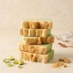 A 300x300 square professional food photograph featuring a tall stack of five thick, rectangular fudge blocks. Each block has a distinct layered pattern: a light, pale green key lime layer and a creamy off-white chocolate layer. The sides and surfaces of each block are heavily coated in crunchy, golden-brown graham cracker crumbs. The stack is centered on a light, textured cream surface. Artfully arranged around the base are small whole key lime wedges and delicate candied lime zest curls. In the foreground, there are loose graham cracker crumbs and two whole graham cracker biscuit pieces. To the right, a small, out-of-focus rose-gold dessert spoon and a rumpled beige linen cloth create a curated, high-end, artisan market display. The lighting is soft and warm, highlighting the fudge's texture and colors.