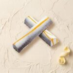 A 300x300 square professional food photograph of two cylindrical gourmet Latte Macchiato truffle bars. The bars are arranged in an "X" shape and feature a sophisticated ombre finish that transitions from creamy white to a deep espresso brown, accented by a bright yellow-gold caramel drizzle running lengthwise. The text "LATTE MACCHIATO" is centered below them in a warm, coffee-toned serif font. The bars rest on a heavily textured, light cream plaster surface. A single, delicate white chocolate curl is placed in the bottom right corner, adding to the artisanal, curated aesthetic. The lighting is soft and bright, highlighting the smooth texture of the chocolate and the artistic details of the garnish.