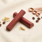 A 300x300 square professional food photograph of two cylindrical gourmet "Manhattan" truffle bars. The bars feature a deep, matte reddish-burgundy hue and are arranged in an "X" shape. The text "MANHATTAN" is centered at the bottom in a classic dark brown serif font. They rest on a soft, light cream-colored linen fabric, providing a gentle texture. The scene is elegantly styled with scattered white chocolate curls, roasted coffee beans, and three polished rose-gold measuring spoons in the upper right corner. The lighting is soft and natural, creating a high-end, sophisticated aesthetic.