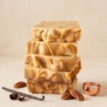 A vertical stack of five smooth and rippled squares of light brown caramel pecan fudge is centered on a textured, light beige surface. The fudge pieces show small embedded pecan bits. Surrounding the base of the stack are loose, whole pecans, a dried vanilla bean pod with delicate dark chocolate curls, and a small copper measuring scoop holding coarse sea salt, all against a textured cream-colored background.