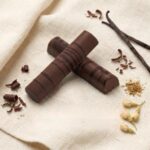 A 300x300 square professional food photograph featuring two cylindrical gourmet "Milk Chocolate Peanut Butter" truffle bars. The bars are made of rich milk chocolate and are decorated with an artistic dark chocolate drizzle pattern. They are arranged in an "X" shape on a soft, cream-colored woven linen surface. The scene is elegantly accessorized with small chocolate curls, whole roasted coffee beans, dried vanilla bean pods, jasmine flower buds, and a dust of edible gold. The text "MILK CHOCOLATE PEANUT BUTTER" is centered at the bottom in a bold, dark brown serif font. The lighting is soft and natural, emphasizing the textures for a high-end, artisanal aesthetic.