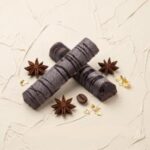 A 300x300 square professional food photograph featuring two cylindrical gourmet "Mocha Madness" truffle bars. The bars have a unique, matte "frosted" dark cocoa finish accented by thin, artistic dark chocolate drizzles. They are arranged in an "X" shape on a neutral, heavily textured cream plaster surface. The scene is elegantly styled with whole roasted coffee beans, three whole star anise pods, and delicate flakes of edible gold leaf scattered around the bars. The text "MOCHA MADNESS" is centered at the bottom in a bold, dark brown serif font. The lighting is soft and natural, creating a sophisticated and high-end artisanal aesthetic.