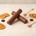 A detailed, premium food photograph of two solid, dark brown, cylindrical chocolate truffle bars. The bars are arranged in a central 'X' shape. The text "OLD FASHIONED" in a dark brown serif font is centered below them. The bars are resting on a hand-textured, light cream plaster or stone surface, replacing the previous plain background. The composition is aesthetically arranged for a curated look, with elements evoking an Old Fashioned cocktail: multiple dried orange wheels (both whole and halved), several whole star anise pods, and two small, polished, twisted copper-handled mixing spoons scattered near the bars. The lighting is soft and natural, emphasizing textures and forms, creating a warm, artisanal feel.
