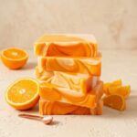A 300x300 square professional food photograph featuring a tall stack of five rectangular fudge blocks. The fudge has a vibrant, marbled pattern of bright citrus orange and creamy white, mimicking an orange creamsicle. The stack is centered on a textured, light cream surface. To the left, a fresh orange is halved, and several orange wedges are scattered to the right. In the bottom-left foreground, a small copper measuring spoon holds coarse sea salt crystals. The background is a soft, warm cream, creating a bright and refreshing aesthetic. Soft lighting highlights the smooth, swirled texture of the fudge.