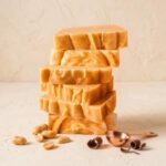 A 300x300 square professional food photograph of a tall stack of six rectangular peanut butter fudge blocks. The fudge is a smooth, golden-tan color with artisanal ridges and soft swirls along the sides. The stack is centered on a light, textured cream-colored surface. In the lower-left foreground, several whole and halved roasted peanuts are scattered. To the right, two dark chocolate curls rest next to a small, polished copper-toned measuring scoop. The background is a minimalist, warm cream, creating a sophisticated and appetizing display. The lighting is soft and natural, emphasizing the creamy texture of the fudge.
