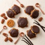 Pecan Caramel Explosions A 300x300 square professional food photograph featuring several "Pecan Caramel Explosions." Each confection consists of a thick swirl of golden caramel sandwiched between roasted pecan halves and topped with a smooth, domed shell of dark chocolate. One central piece is decorated with a flake of edible gold leaf and a pinch of sea salt. The treats are arranged on a neutral, textured cream plaster surface. The scene is artistically styled with scattered whole pecans, dried vanilla bean pods, and several polished copper-toned measuring spoons. The lighting is soft and bright, creating a high-end, artisanal aesthetic.