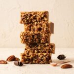A 300x300 square professional food photograph of a tall stack of five rectangular fudge blocks. The front of each slice is densely packed with a mix of large, whole toasted pecan halves and smaller chopped pecan pieces, pressed into a golden-brown fudge base. The stack is centered on a light, textured cream-colored surface. In the foreground, several whole pecans and dark chocolate curls are scattered around the base. To the right, a small copper-toned measuring scoop holds a pinch of coarse white sea salt. The background is a minimalist, warm cream, creating an artisanal and high-end aesthetic. Soft, natural lighting highlights the crunchy texture of the pecans and the smooth fudge sides.