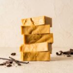 A 300x300 square professional food photograph of a tall stack of five rectangular Penuche fudge blocks. The fudge is a uniform, warm brown sugar and caramel color with a matte, creamy finish and subtle artisanal ripples on the sides. The stack is centered on a light, textured cream-colored surface. In the lower-left, several dark vanilla bean pods and a few small chocolate shavings are artfully arranged. To the right, additional dark chocolate curls rest near the base. The background is a minimalist, textured cream color, creating a warm, sophisticated aesthetic. Soft lighting highlights the dense, smooth texture of the brown sugar fudge.