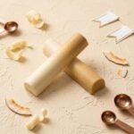 A 300x300 square professional food photograph featuring two cylindrical gourmet Piña Colada truffle bars. The bars are arranged in an "X" shape and feature a beautiful ombre finish, transitioning from a creamy white to a warm golden-yellow. The text "PINA COLADA" is centered at the bottom in a bold, gold-toned serif font. The bars rest on a textured, light cream-colored surface. Scattered around the base are small dried coconut pieces, whole roasted cacao nibs, elegant curved white chocolate shavings, and two polished copper-toned measuring spoons. The lighting is soft and natural, creating a high-end, artisanal aesthetic.