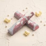 A 300x300 square professional food photograph featuring two cylindrical gourmet Pink Champagne truffle bars. The bars are arranged in an "X" shape and feature an artisanal marbled finish in shades of dusty rose, soft grey, and creamy white, accented with fine white speckles. The text "PINK CHAMPAGNE" is printed twice at the bottom in a sophisticated mauve-toned serif font. The bars rest on a textured, light cream-colored surface. The composition is elegantly decorated with white chocolate curls, delicate white decorative pearls, and tiny silver sugar crystals. The lighting is soft and romantic, creating a high-end, celebratory, and artisanal aesthetic.