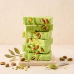 A 300x300 square image featuring a stack of six thick, vibrant green pistachio fudge bars. Whole and crushed roasted pistachios are embedded throughout and scattered across the surface of each slice. The stack sits on a small marble platform against a soft, textured cream background. Surrounding the base are whole pistachios, a sprig of green leaves, and a decorative ceramic spoon filled with fine green pistachio powder. Delicate gold flakes are scattered around the scene, enhancing the premium, artisanal feel.