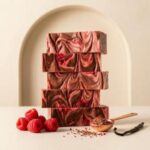 A 300x300 square professional food photograph featuring a tall stack of five thick fudge slices. The fudge has an elegant, marbled swirl of deep cocoa brown and vibrant raspberry pink. The stack is centered on a light cream surface against a soft, arched background. At the base, a cluster of fresh, ripe red raspberries sits to the left. To the right, a rose-gold measuring spoon is filled with chocolate shavings and dried raspberry bits, accompanied by a single vanilla bean pod. The lighting is soft and natural, emphasizing the rich, creamy texture of the artisanal confection.