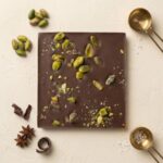 A 300x300 square professional flat-lay photograph featuring a square Belgian milk chocolate bar. The surface of the chocolate is studded with whole and crushed vibrant green roasted pistachios. The bar is centered on a light, textured cream-colored surface. To the left, small clusters of whole pistachios, dark chocolate curls, and star anise are artfully arranged. To the right, two elegant gold-toned measuring scoops rest on the surface, one containing coarse sea salt crystals. The lighting is soft and natural, emphasizing the rich, smooth texture of the chocolate and the crunchy, colorful pistachio topping for a sophisticated, artisanal look.