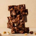 A 300x300 square professional food photograph featuring a tall, slightly uneven stack of five thick chocolate rocky road fudge slices. The fudge is deep brown and densely packed with visible mini marshmallows and roasted peanuts, creating a rugged, artisanal texture. The stack is centered on a textured cream surface. Artfully arranged at the base are a toasted marshmallow, a few whole roasted peanuts, and a delicate dark chocolate curl. A rose-gold spoon handle is partially visible in the bottom-left corner. The background is a soft, warm cream, giving the image a clean and high-end aesthetic.